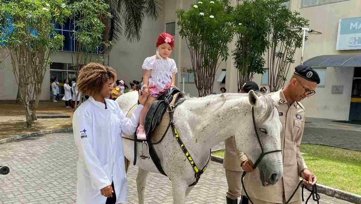 Criança, paciente do HEC, andando a cavalo, guiada por policial, com ajuda de colaboradora do hospital