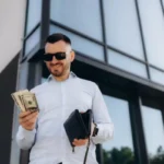 jovem-homem-de-negocios-de-pe-apoiado-na-parede-usando-oculos-na-rua-da-cidade-contando-dinheiro-sorrindo-alegremente_633611-10365